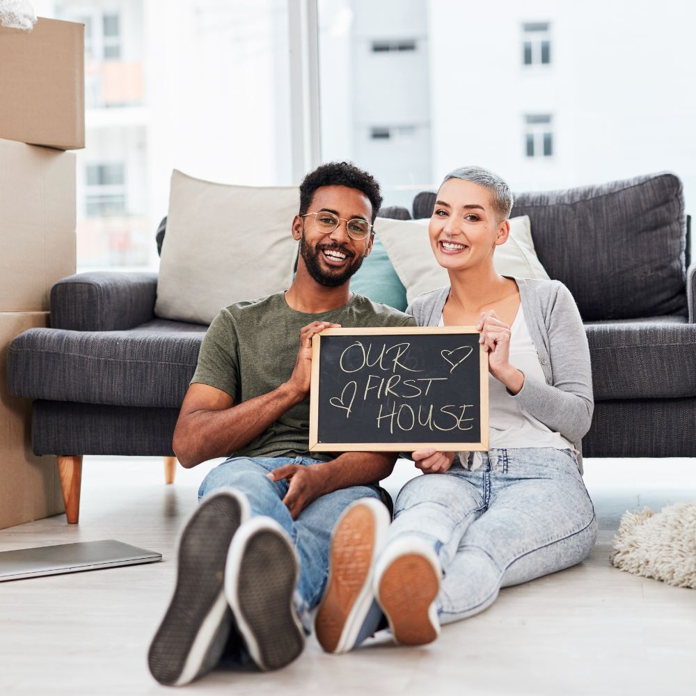 A couple (black male, white female) sat on the floor in front of their sofa, holding a blackboard that states 'our first house' bought during a volatile mortgage market