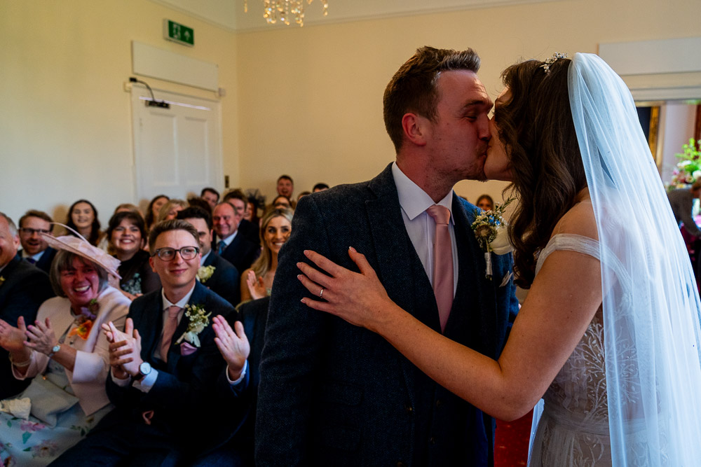 The bride and groom kiss in front of their guests after finishing their vows. The picture is quite dark
