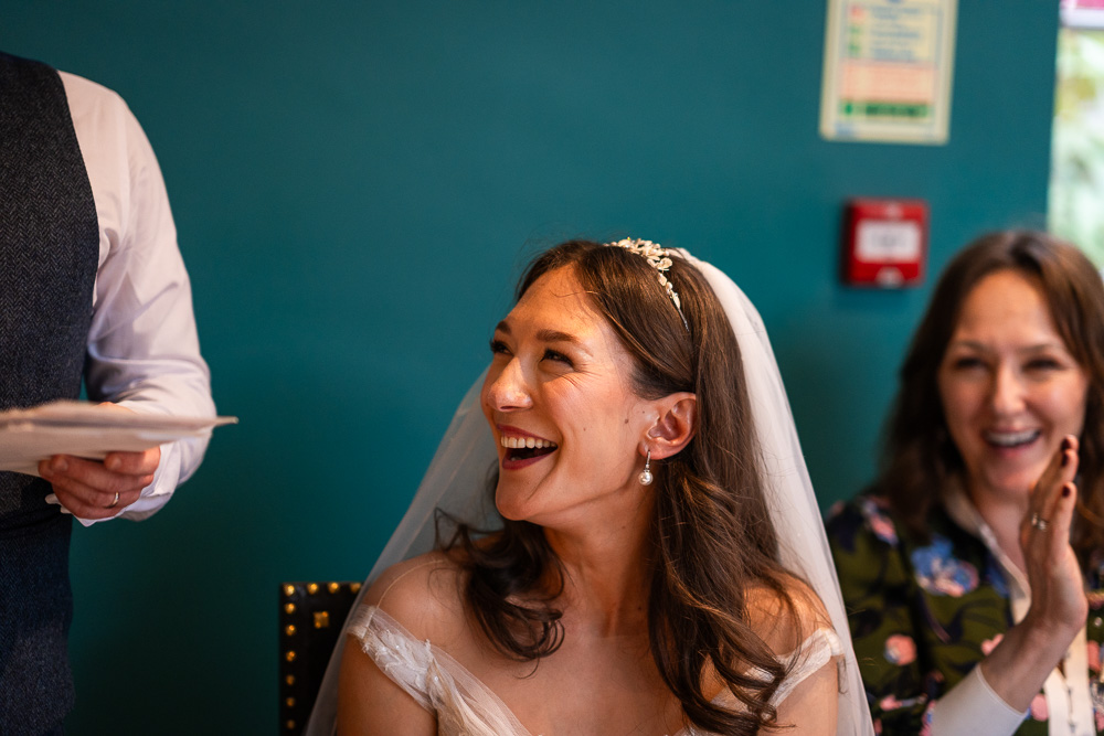 Prices of wedding photography before an image showing the bride laughing with the groom's arm to the left and a fire alarm and signage on the wall behind her.