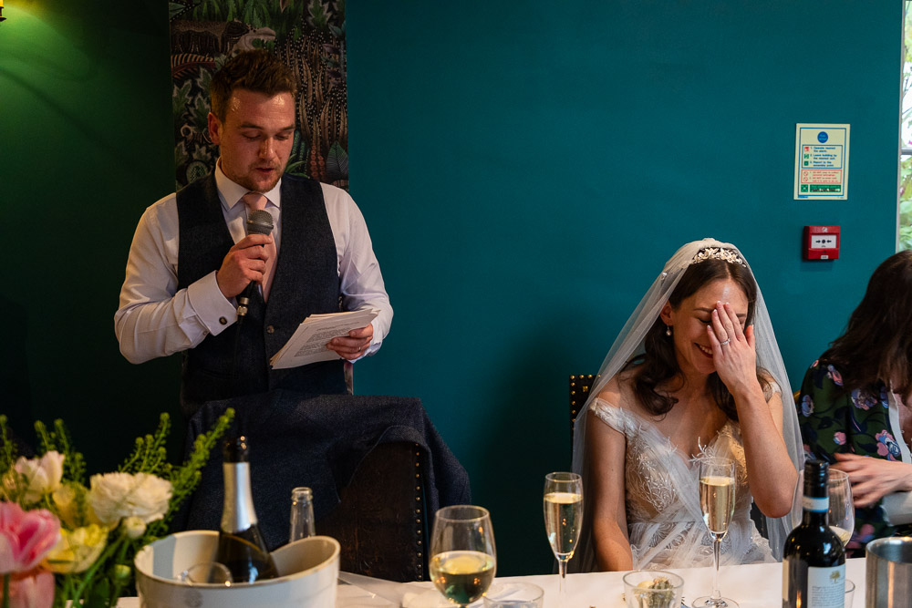 prices of wedding photography - a groom making his speech into a microphone as his bride holds a hand to her face, laughing. There's a fire alarm and a sign on the wall behind her, and it is quite a dark image