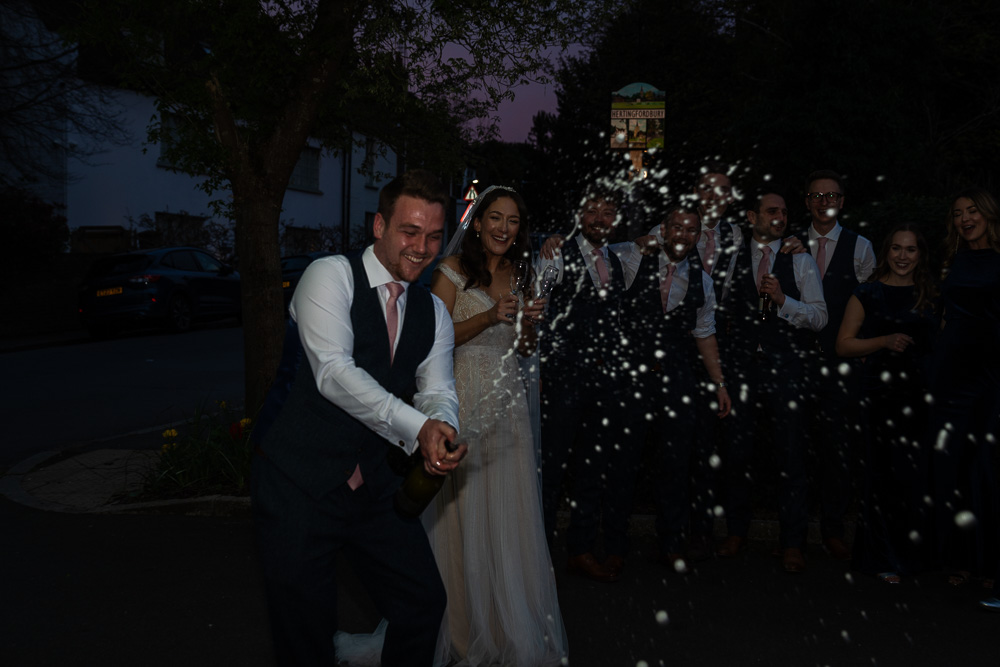 Very dark image of a groom spraying champagne with his bride and groomsmen in the background, barely visible
